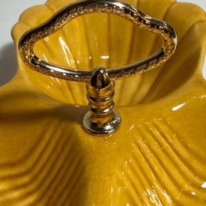 May include: A mustard yellow ceramic serving dish shaped like a shell with a glossy crackle glaze finish. The dish features a central gold-toned metal handle with an ornate, textured pattern and a tiered, pedestal-style base attachment.