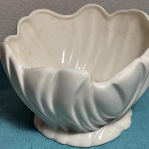 May include: A glossy off-white ceramic bowl featuring a scalloped rim and a molded, fluted exterior design that mimics the appearance of a shell. The bowl has a wide, open top and a circular base with a matching scalloped edge detail.