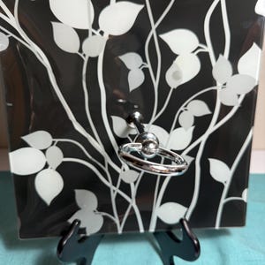 May include: A square glass plate featuring a black background with a white botanical leaf and vine pattern. The plate has a central chrome metal post with a circular ring handle attached to the front, supported by a black plastic stand.