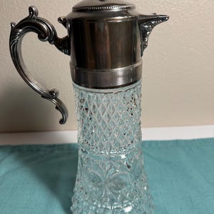 May include: A clear glass carafe with a diamond-cut pattern and a silver-toned metal lid and handle. The metal top features a pointed finial, a spout, and a ribbed band where it connects to the glass body. The handle has a curved, ornate shape.