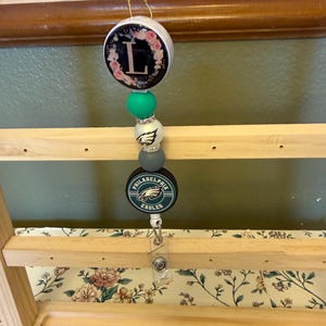 Puede incluir: Un carrete de insignia con cuentas con un diseño floral de la inicial "L", cuentas verdes y blancas, y un logotipo de los Philadelphia Eagles. El carrete tiene un clip de plástico transparente para sujetarlo a la ropa.