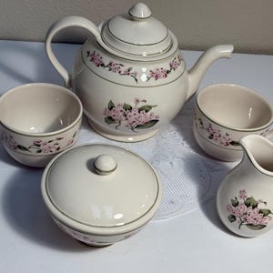 May include: A cream-coloured ceramic tea set featuring a teapot with a lid, a lidded sugar bowl, a creamer, and two tea cups. Each piece is decorated with a delicate pattern of pink floral clusters and green leaves, accented by thin sage green trim.