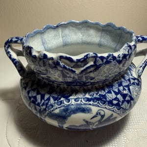 May include: A ceramic bowl with two handles, featuring a white base and intricate blue patterns. The design includes depictions of birds and trees, with a scalloped rim and a geometric border around the upper section of the rounded vessel.