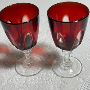 May include: A pair of matching stemmed glasses featuring deep red bowls with vertical fluted detailing. Each glass has a clear, faceted glass stem and a circular clear glass base, providing a stable foundation for the vibrant red upper portion.