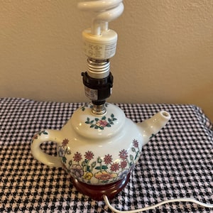 Könnte beinhalten: Eine weiße Teekannen-Lampenbasis aus Keramik mit einem bunten Blumenmuster aus rosa, gelben und violetten Blüten sowie grünen Blättern. Die Lampe hat eine schwarze Fassung und eine weiße spiralförmige Glühbirne an der Oberseite.