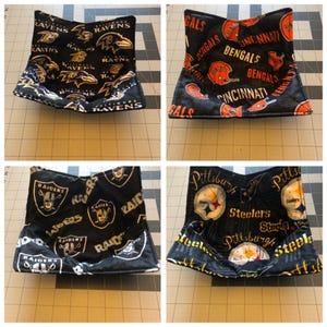 Pode incluir: Quatro forros de tigela de tecido preto com diferentes logotipos de times da NFL. Os logotipos são Baltimore Ravens, Cincinnati Bengals, Las Vegas Raiders e Pittsburgh Steelers.