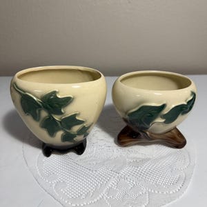 May include: Two cream-coloured ceramic planters with a glossy finish. Each planter features a raised, dark green ivy leaf design on the exterior. The left planter has a dark brown base, while the right planter sits on a brown, branch-like base.