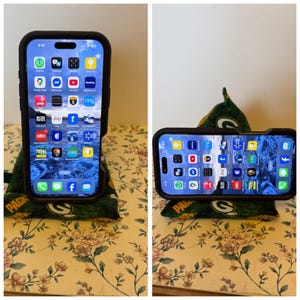 Könnte beinhalten: Ein schwarzes Smartphone in einer Schutzhülle wird auf einem grün-goldenen Handyständer aus einem Sitzsack präsentiert. Der Telefonbildschirm zeigt App-Symbole. Auf dem Sitzsack steht das Wort "PACK".