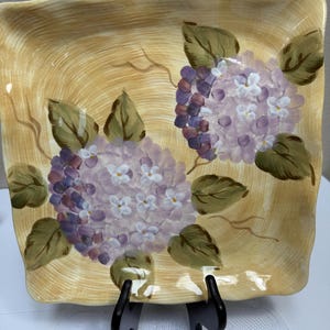 May include: A square ceramic plate with a wavy edge featuring a hand-painted tan background with brushstroke textures. The plate displays two clusters of purple and white hydrangea flowers with green leaves and thin brown stems painted on the surface.