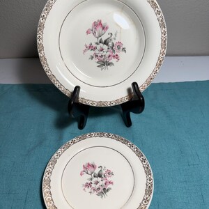 May include: Two cream-colored ceramic plates featuring a central floral bouquet with pink and grey flowers. The rims are decorated with an intricate gold filigree pattern. The larger plate is displayed on a black stand and shows a visible crack.