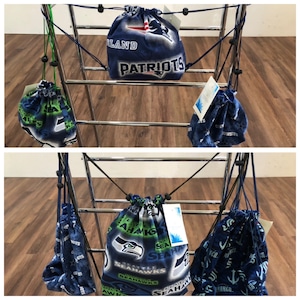 May include: A metal rack with six drawstring bags hanging from it. The bags are made of fabric and feature different sports team logos, including the New England Patriots, Seattle Seahawks, and the Green Bay Packers.