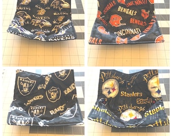 May include: A set of four fabric bowl cozies featuring NFL team logos and names. The designs include the Baltimore Ravens, Cincinnati Bengals, Las Vegas Raiders, and Pittsburgh Steelers, all printed on black fabric with team-specific colours.
