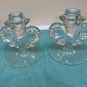 Puede incluir: Un par de candelabros de vidrio transparente con un soporte cilíndrico central, un tallo estriado y dos asas simétricas en forma de voluta a los lados. La base circular de cada candelabro presenta un patrón floral grabado.