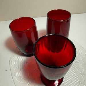 Könnte beinhalten: Drei rubinrote Glastrinkbecher mit glatter, zylindrischer Form. Das vorderste Glas weist vertikale Rillendetails an den Innenwänden auf, während die beiden dahinter positionierten Gläser eine schlichte, glatte Außenfläche besitzen.