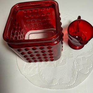 May include: A square red glass canister with a hobnail texture on the exterior and a matching red glass creamer pitcher with a handle and spout. Both items are made of translucent red glass and feature a distinct raised dot pattern on the surface.