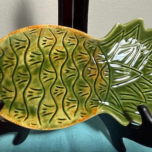 May include: A ceramic pineapple-shaped serving dish featuring a glossy green glaze with textured, embossed scale patterns and orange-toned edges. The piece has a distinct leafy top section and a rounded base, finished with a smooth, reflective surface.