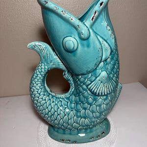 May include: A turquoise ceramic fish-shaped pitcher with a glossy crackle glaze finish. The pitcher features a wide, open-mouthed top, detailed scale textures, and a curved tail handle. Distressed brown edges accent the fins, rim, and base.