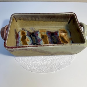 Puede incluir: Una fuente de horno rectangular de cerámica con dos asas laterales. El exterior y el interior presentan un esmalte moteado en tonos beige, marrón y rojo intenso, con un borde de color marrón rojizo más oscuro.