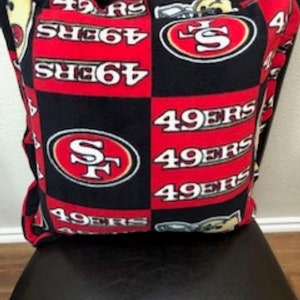 May include: A red, black, and white fleece pillow with the San Francisco 49ers logo and the words "49ers" repeated on it.