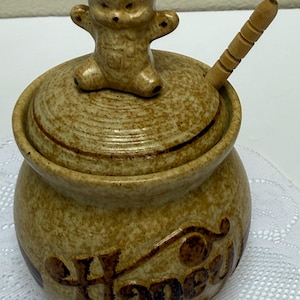 May include: A speckled tan ceramic honey pot with a rounded body and a matching lid featuring a sculpted bear figurine on top. The word Honey is embossed in dark brown script on the front of the pot, which includes a wooden honey dipper.