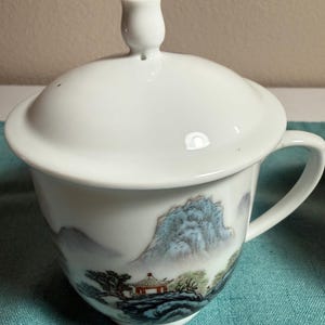 May include: A white porcelain tea cup with a matching lid featuring a knob handle. The exterior surface is decorated with a painted landscape scene depicting blue mountains, green trees, and a small structure with a red roof near the base.