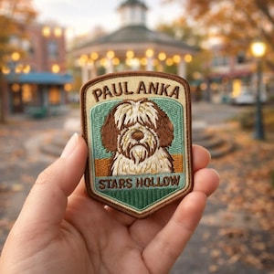 May include: Embroidered patch featuring a dog with the text "PAUL ANKA" and "STARS HOLLOW". The patch has a tan border, a green and blue background, and a brown and white dog illustration. The patch is held in a hand.