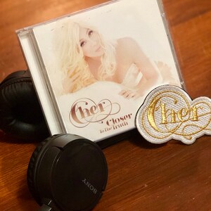 May include: A Cher Closer to the Truth CD album case featuring a portrait of Cher, black Sony headphones, and a white embroidered patch with the gold-colored text Cher in a cursive script, all displayed together as a collection of music memorabilia.
