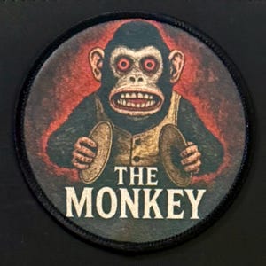 May include: Circular patch featuring a cartoon monkey with red eyes, holding cymbals. The monkey wears a vest and has a wide grin. The text "THE MONKEY" is below the monkey. The patch has a black border and a red background.