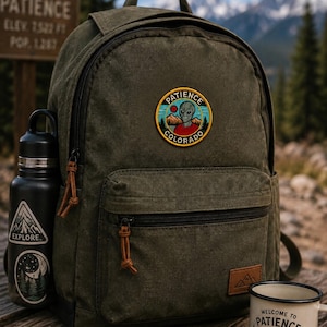 May include: An olive green canvas backpack featuring a circular patch with an alien graphic and the text PATIENCE COLORADO. A black water bottle with mountain and moon stickers sits on the left, and a white enamel mug with mountain art is nearby.