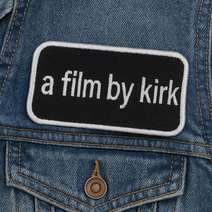 May include: A denim jacket with a black rectangular patch that reads "a film by kirk" in white text. The patch is bordered with a white outline. The jacket has a chest pocket with a button.