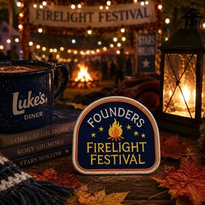 May include: A navy blue embroidered patch with a white border featuring a central campfire graphic, yellow stars, and the text FOUNDERS FIRELIGHT FESTIVAL in yellow lettering, displayed alongside a blue Luke's Diner mug and stacked books.