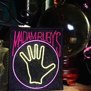 May include: A black embroidered patch with the words "MADAM RUBY'S" in pink, and a yellow hand design inside a pink circle. The patch is rectangular and appears to be made of fabric.
