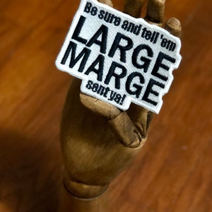 May include: A rectangular embroidered patch with a white background and a black stitched border. The patch features the black embroidered text "Be sure and tell 'em LARGE MARGE sent ya!" in a bold, sans-serif font style across the surface.