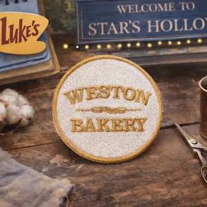 May include: A round Weston Bakery patch with gold lettering and trim sits on a wooden surface, next to a steaming copper mug of coffee and a pair of decorative scissors. A sign that says "Welcome to Star's Hollow" is in the background.
