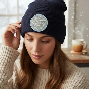 May include: Navy blue beanie with a circular patch that reads "I Smell Snow" in a whimsical font. The patch features snowflakes and stars in light blue and yellow. The beanie is a winter accessory.