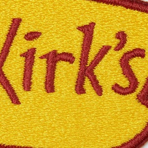 May include: Embroidered patch with the text "Kirk's" in a bold, red, stitched font. The patch has a yellow background and a red border, creating a striking contrast. The texture of the embroidery is visible, adding depth to the design.