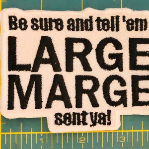 K&ouml;nnte beinhalten: Ein rechteckiger, wei&szlig;er, bestickter Aufn&auml;her mit schwarzem Text, der "Be sure and tell 'em LARGE MARGE sent ya!" lautet. Der Aufn&auml;her liegt auf einer gr&uuml;nen Schneidematte mit gelbem Raster und ist etwa 9 cm breit.