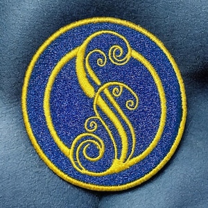 May include: A blue and gold embroidered patch with a stylized design of a wave or water.
