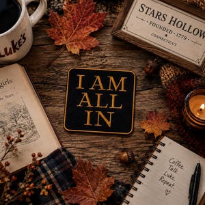 May include: A black square patch with gold embroidered text reading I AM ALL IN, surrounded by a white mug with Luke's text, a book titled Gilmore girls, a Stars Hollow sign, a notebook with Coffee. Talk. Luke. Repeat. text, autumn leaves, and a candle.