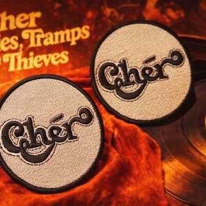 May include: Two circular patches with the name "Chér" embroidered in black on a light beige background. The patches are set against a backdrop of a vintage album cover and vinyl records, with the text "Cher, Gypsies, Tramps & Thieves".