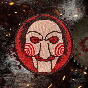 May include: Embroidered patch featuring a red and black circular design of a horror movie character with a red spiral pattern on the cheeks and a wide, toothy grin.
