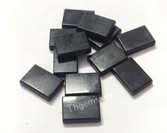 May include: A collection of small, rectangular, dark gray gemstones scattered on a white surface. The gemstones have a polished, reflective surface. The text "Thgemskey" is visible on one of the gemstones.