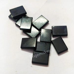 以下が含まれることがあります： 小さく長方形で研磨された黒い石のコレクション。石は白い表面に散らばっており、一部は重なっています。石の表面は光を反射し、ハイライトを作り出しています。「Tngemskey」というテキストが表示されています。