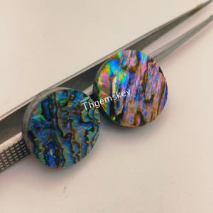 May include: Two round, iridescent abalone shell cabochons with a mix of blue, green, and brown hues. The cabochons are held by a pair of silver tweezers. The text "Thgemskey" is visible on the cabochons.
