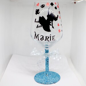 May include: A clear glass wine glass with a blue glitter stem and base. The glass features a black silhouette of Alice in Wonderland falling down a rabbit hole, surrounded by playing card symbols. The name "Marie" is written in black lettering on the glass.