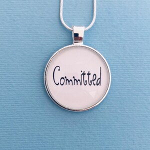 May include: A silver-toned necklace with a round pendant. The pendant has a white background with the word "Committed" written in black cursive script. The necklace is displayed against a light blue background.