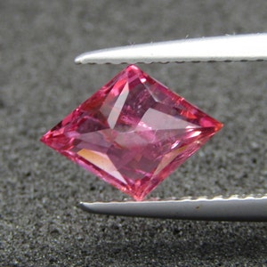 May include: A pink gemstone with a square cut, held by tweezers. The gemstone is clear and has a slight shimmer.