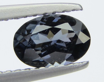 May include: A dark gray oval-shaped gemstone with facets, held by tweezers.