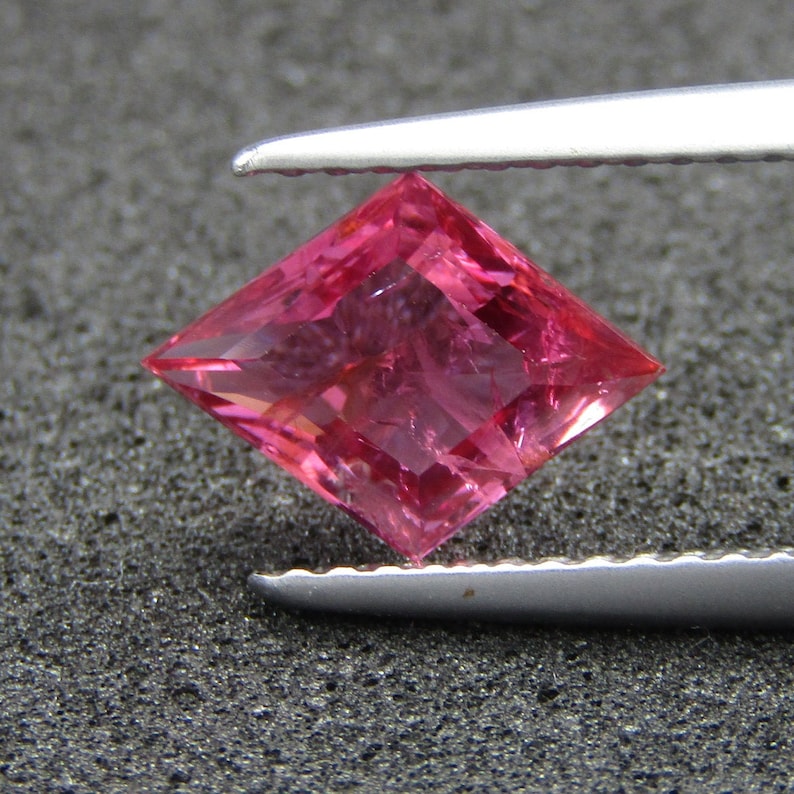 May include: A pink gemstone with a square cut, held by tweezers against a dark gray background.