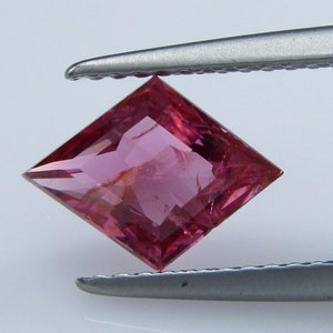 May include: A pink square-shaped gemstone with a faceted cut. The gemstone is held by tweezers.